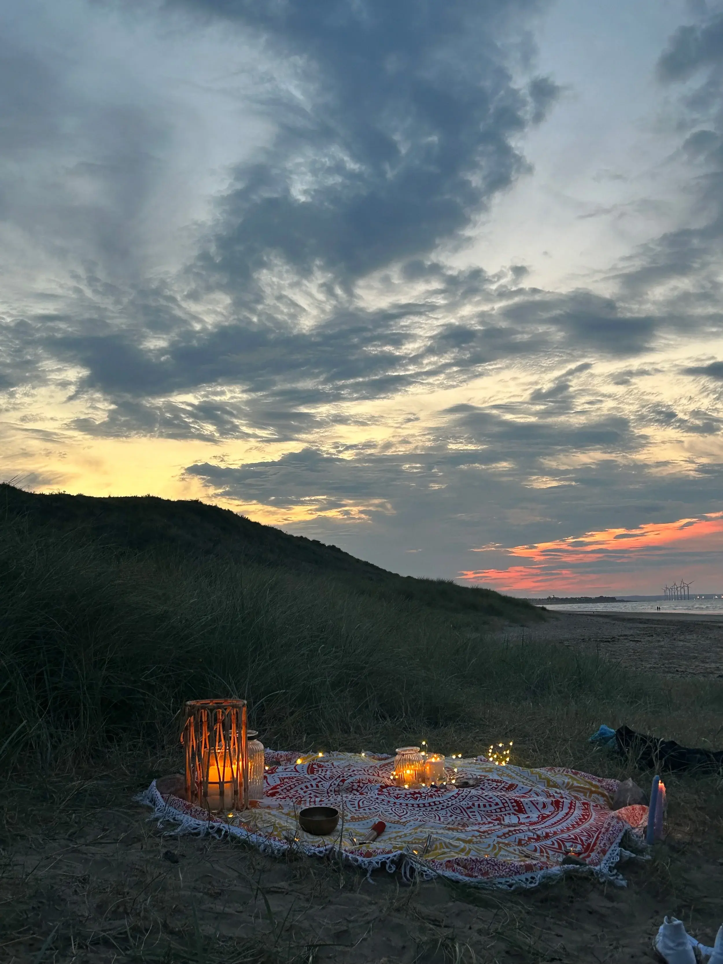 Candles and fairy lights laid out on a blanket on the dunes at dusk, the sea and a pink sunset beyond